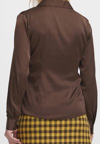 Blusa lunga in raso marrone con colletto, caratterizzata da una texture liscia e un design aderente, abbinata a una gonna a scacchi gialla e nera.