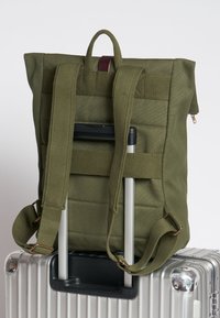 Mochila de lona verde oliva con correas ajustables, diseño acolchado y un asa superior; montada en el mango de una maleta plateada.