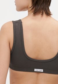 Sivý bavlnený bralet s širokými ramienkami, zaobleným výstrihom a hladkou textúrou. Má štítok s názvom značky na chrbte.