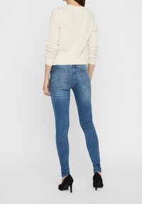 Blusa de manga larga crema y jeans ajustados azules vistos desde la parte posterior, con dos bolsillos traseros y tacones altos, sobre un fondo blanco liso.