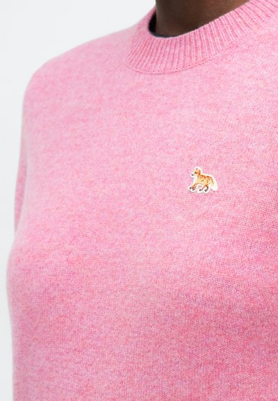 Zbliżenie na różowy sweter z dzianiny z małym haftowanym pomarańczowym emblematem lisa po lewej stronie klatki piersiowej.