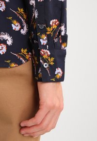 Granatowa koszula w kwiaty z mankietami na guziki; ma kolorowe wzory, miękką teksturę oraz dopasowany krój z lekko zaokrąglonym dołem.