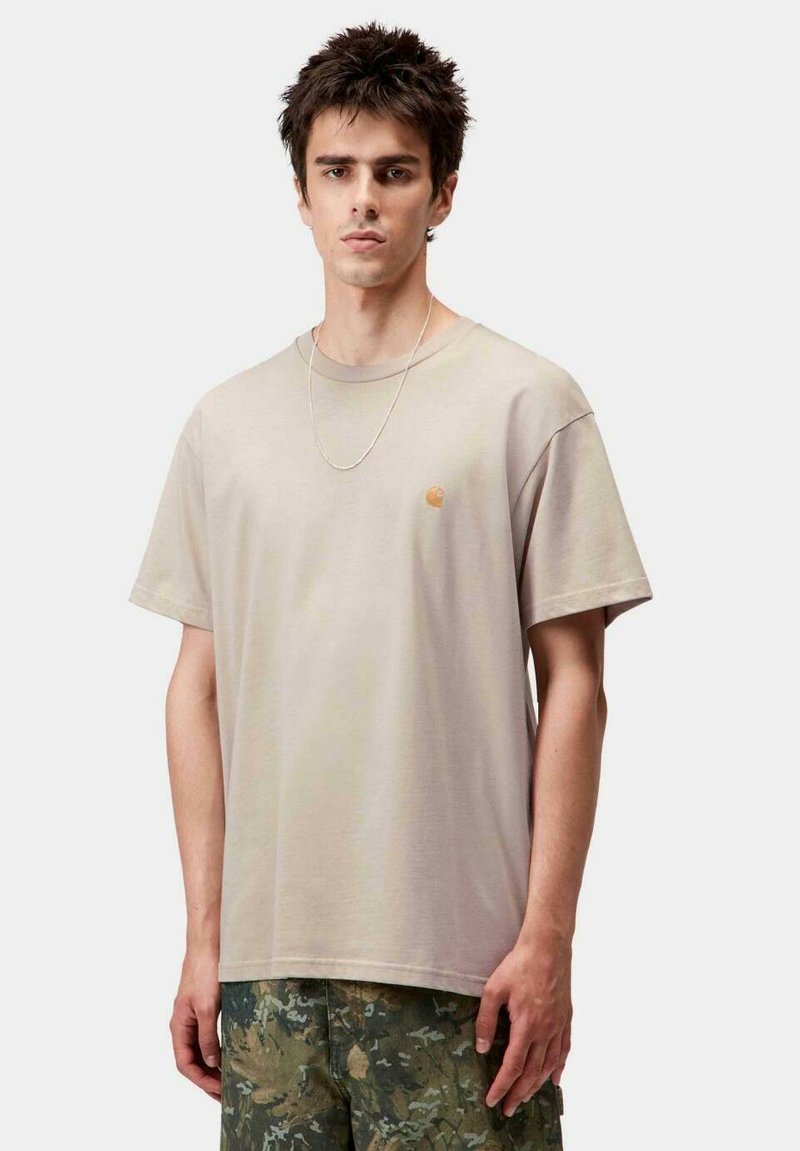 Camiseta de algodón beige con cuello redondo y mangas cortas. Presenta un pequeño logotipo naranja en el lado izquierdo del pecho. Combinada con pantalones cortos de camuflaje.