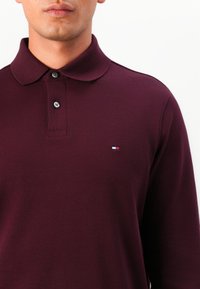 Burgundy long-sleeve polo shirt made of textured fabric, featuring a classic collar, two button placket, and a small logo on the chest.