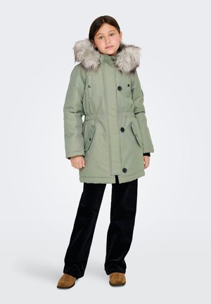 Grön vadderad parka med en huva av syntetisk päls, med dragsko i midjan och knäppningar, ihop med svarta kordflorsbyxor och bruna skor.