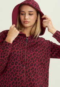 Sudadera con cremallera en color burdeos con estampado de leopardo negro. Cuenta con capucha con cordón, mangas largas y textura de tela suave.