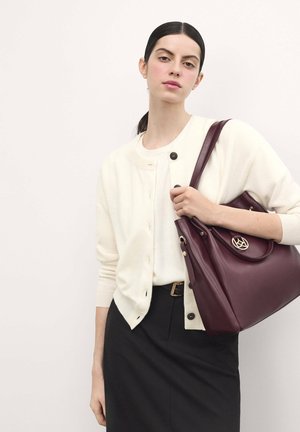 Cardigan bianco con bottoni neri, indossato su una maglietta crema. Borsa in pelle bordeaux con hardware dorato, caratterizzata da un accento con logo circolare.