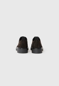 Zapatos de ante negro con un perfil bajo, que presentan una superficie texturizada y suelas de goma. La vista trasera muestra detalles elásticos y un logotipo.