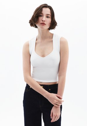 Débardeur blanc court avec un décolleté arrondi et de larges bretelles, associé à un jean bleu foncéOr avec des détails en métal doré.