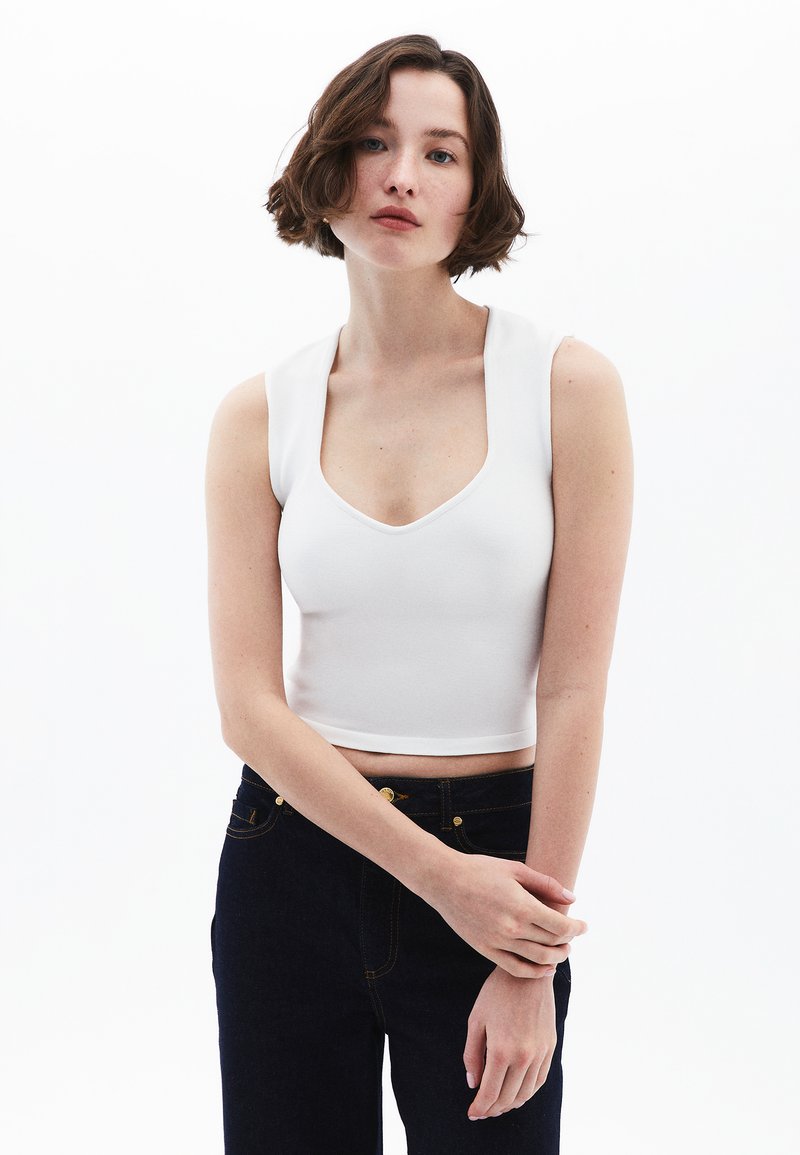 Top corto bianco con scollo rotondo e spalline larghe, abbinato a jeans blu scuro con dettagli in hardware dorato.