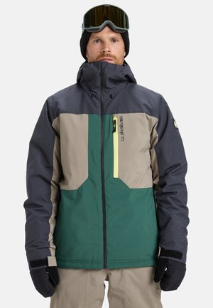 Homme portant une veste de ski multicolore, un pantalon beige, des gants noirs, un bonnet noir et des lunettes de ski sur le front, se tenant devant un fond blanc.