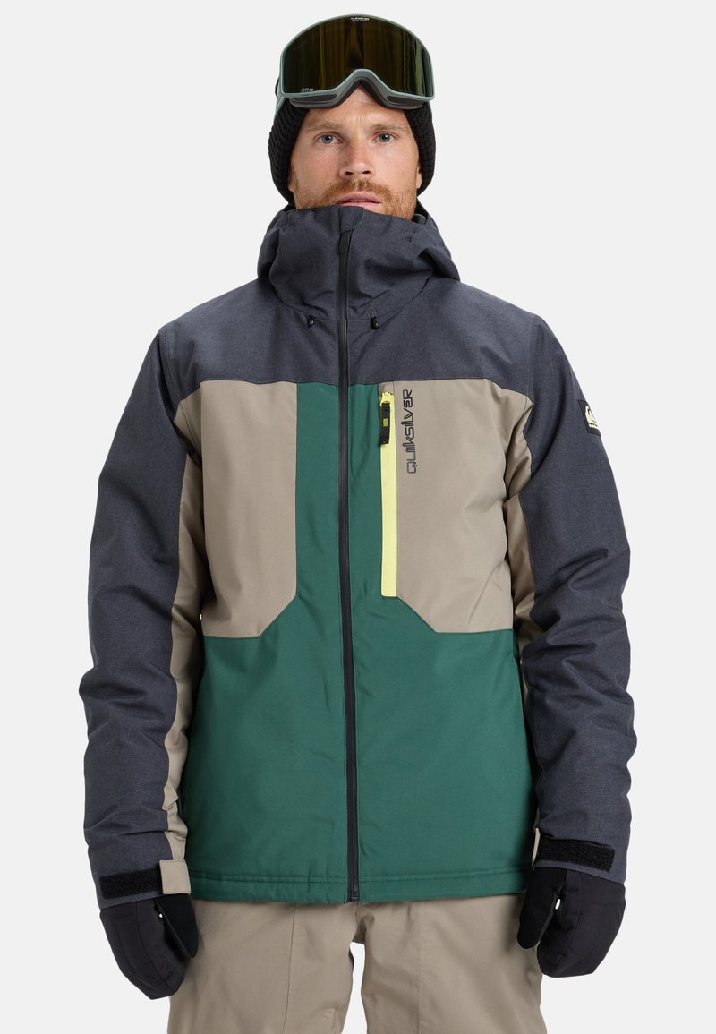 Mann in einer mehrfarbigen Skijacke, beigen Hosen, schwarzen Handschuhen, einer schwarzen Mütze und Skibrille auf der Stirn, der vor einem weißen Hintergrund steht.