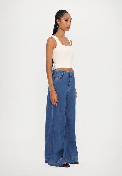 Wide-leg blue denim jeans with a high waist, featuring a button closure and front pockets. Worn with a fitted cream tank top.