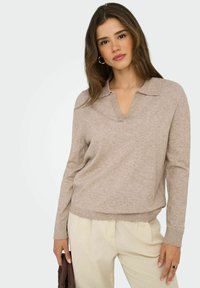 Mujer con cabello largo castaño vistiendo un suéter beige de escote en V y pantalones de color claro, sosteniendo un bolso marrón doblado al lado.