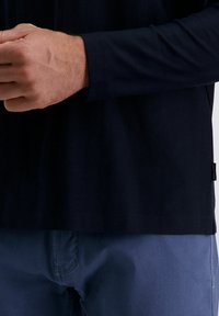 Marineblaues Langarmhemd mit Fischgrätmuster, gerade geschnittenem Saum und einem Etikett an der Seite; kombiniert mit blauen Hosen und sichtbaren Nähten.