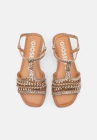 Sandalias con tiras y acentos trenzados metálicos, suela de imitación cuero en tono marrón, y tacón plano. La parte superior presenta tiras horizontales y verticales.