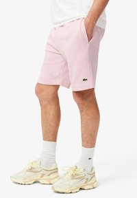 Shorts de algodón rosa claro con bolsillos laterales, con un logo verde de Lacoste en el dobladillo. Se llevan con zapatillas beige y calcetines blancos.