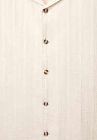 Gros plan d'une chemise en tissu texturé crème avec cinq boutons ronds marron alignés verticalement au centre.