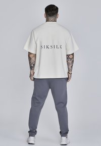 Kremowa koszulka z krótkim rękawem z logo "SIKSILK" na plecach, sparowana z szarymi joggerami. Posiada luźny krój i miękką teksturę materiału.