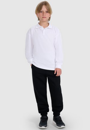 Niño rubio de pie con una camisa polo blanca de manga larga, pantalones negros y zapatillas negras contra un fondo liso y claro.