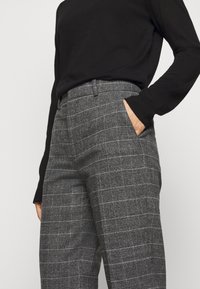 Un pantalon à carreaux gris avec une coupe ajustée, à devant plat et poches latérales, fabriqué dans un tissu texturé. Assorti à un haut noir à manches longues.