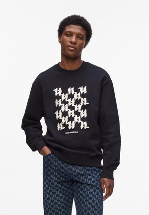 Sort sweatshirts med et gentaget hvidt grafisk mønster og teksten "KARL LAGERFELD" nederst. Kombineret med mørke mønstrede jeans.