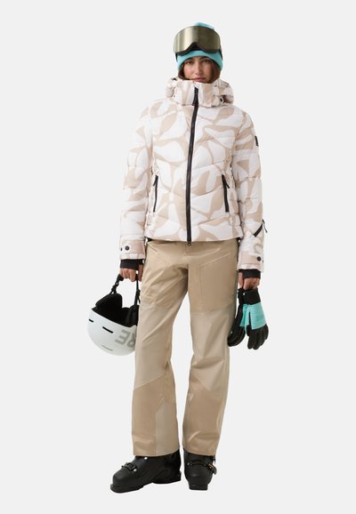 Skijakke med et abstrakt mønster i hvid og beige, kombineret med beige bukser, sorte skistøvler, grønne og sorte handsker samt en hvid hjelm.