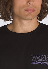 Camiseta de algodón negra con cuello redondo que presenta el logo púrpura "VANS Design Co. custom classics" en el pecho. Textura suave y ajuste informal.