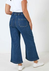 Blaue Denim-weit geschnittene Hose mit elastischem Bund, ausgestattet mit zwei Gesäßtaschen und einem rohen Saum. Getragen mit weißen Sneakers.