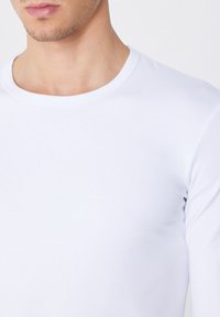 Camiseta de manga larga blanca con cuello redondo, hecha de una tela suave y elástica. Presenta un diseño ajustado con costuras mínimas y un escote clásico.