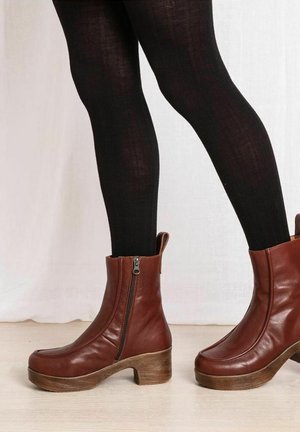 Brown leather ankle boots with a chunky wooden heel, side zipper, and pull tab, paired with black leggings, against a light backdrop.