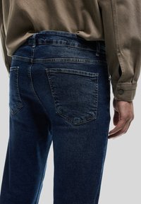 Tummansiniset farkut, joissa on klassinen viisi-taskuinen muotoilu. Sileä pinta ja suora lahje, näkyvät tikkaukset sekä läpälliset takataskut.