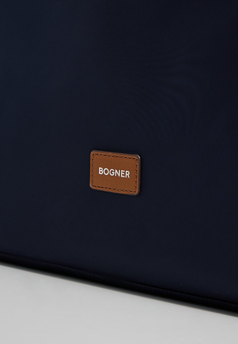 Marineblaues Täschchen mit glatter Textur, versehen mit einem rechteckigen, braunen Lederschild, auf dem "BOGNER" in weißer Schrift geprägt ist.
