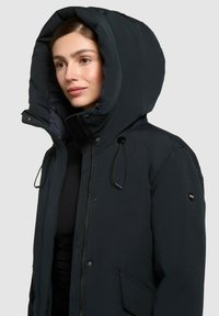 Femeie îmbrăcată cu o jachetă de iarnă neagră cu glugă, fermoar și butoane, stând pe un fundal gri deschis uniform.