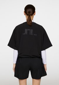 Musta ylisuuri t-paita lyhyillä hihoilla, jossa on pisteellinen geometrinen kuvio selässä. Materiaali vaikuttaa sileältä ja pehmeältä.