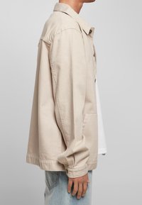 Mężczyzna ubrany w beżową kurtkę z guzikami na białą koszulę i jasnoniebieskie dżinsy, stojący bokiem na jednolitym szarym tle.