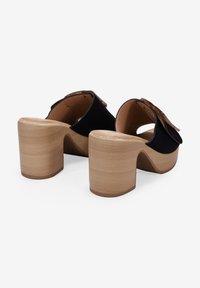 Zuecos de ante negro con la parte trasera abierta, un ancho tacón de madera y una correa decorativa en la parte superior. Acabado suave, diseño sencillo.