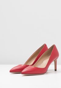 Une paire d'escarpins rouges en cuir verni avec des pointes à talons hauts et des semelles intérieures beiges sur une surface blanche, contre un fond gris uni.