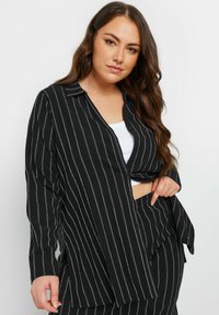 Camicia a righe bianche e nere con una vestibilità comoda, maniche lunghe, colletto e spacco laterale. Il tessuto appare strutturato con una finitura liscia.