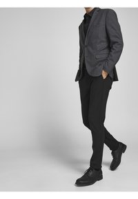 Grijze blazer met patroon over een zwarte shirt, gecombineerd met zwarte broek en leren nette schoenen. De blazer heeft gestructureerde revers en zakken.