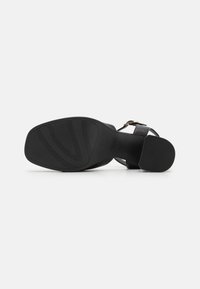 Svart hæl sandal med en glatt skinnoverdel og en teksturert gummisåle. Har en gullfarget spenne og design med krossrem.