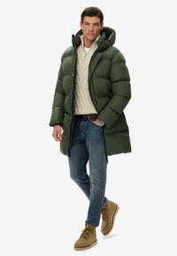 Mężczyzna w zielonej kurtce puchowej z kapturem, beżowym swetrze w warkocze, niebieskich dżinsach, brązowych butach i zegarku na nadgarstku, stojący z jedną ręką w kieszeni.