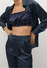 Frau in dunkelblauen gemusterten Hosen und passender offener Bluse über einem marineblauen floralen Crop-Top, die Hände in die Hüften gestützt.