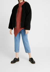 Giacca bomber nera e morbida su un maglione rosso a treccia con parete asimmetrica, abbinata a jeans cropped blu chiaro e scarpe nere.