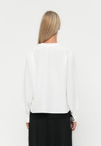 Witte lange mouwen shirt met een ronde halslijn en geplooide achterkant. De stof lijkt lichtgewicht en lichtjes gestructureerd.