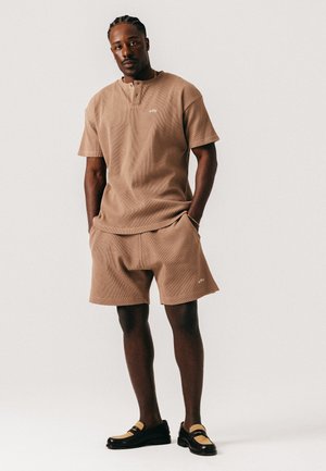 Mand med flettet hår iført tan-farvet kortærmet skjorte og matchende shorts med sorte og tan loafers, stående mod en hvid baggrund.