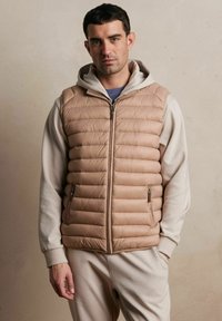 Beżowa kamizelka ocieplana z poziomym przeszyciem i kieszeniami na zamek, noszona na jasnoszarym kapturem. Gładki, miękki materiał w luźnym fasonie.