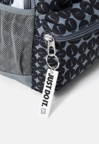 Σακίδιο Nike με γκρίζο και μαύρο γεωμετρικό μοτίβο, πλέγμα πορτοκάλι πλευρική τσέπη, ασημένιο φερμουάρ με ετικέτα μπρελόκ που γράφει "JUST DO IT."