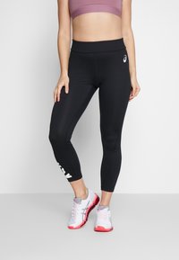 Czarne legginsy sportowe o dopasowanym kroju i wysokim stanem, z białym logo na dolnej części nogawki, w połączeniu z jasnoszarymi trampkami.