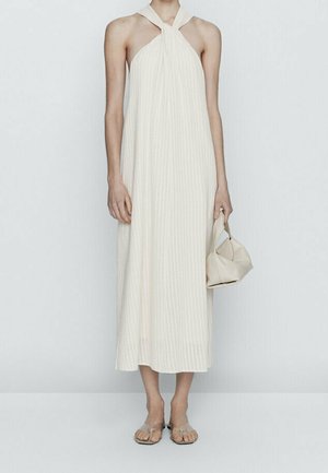 Robe midi à col licou, beige clair avec des rayures verticales. Portée avec des sandales plates. Le modèle tient un petit sac beige à texture douce.